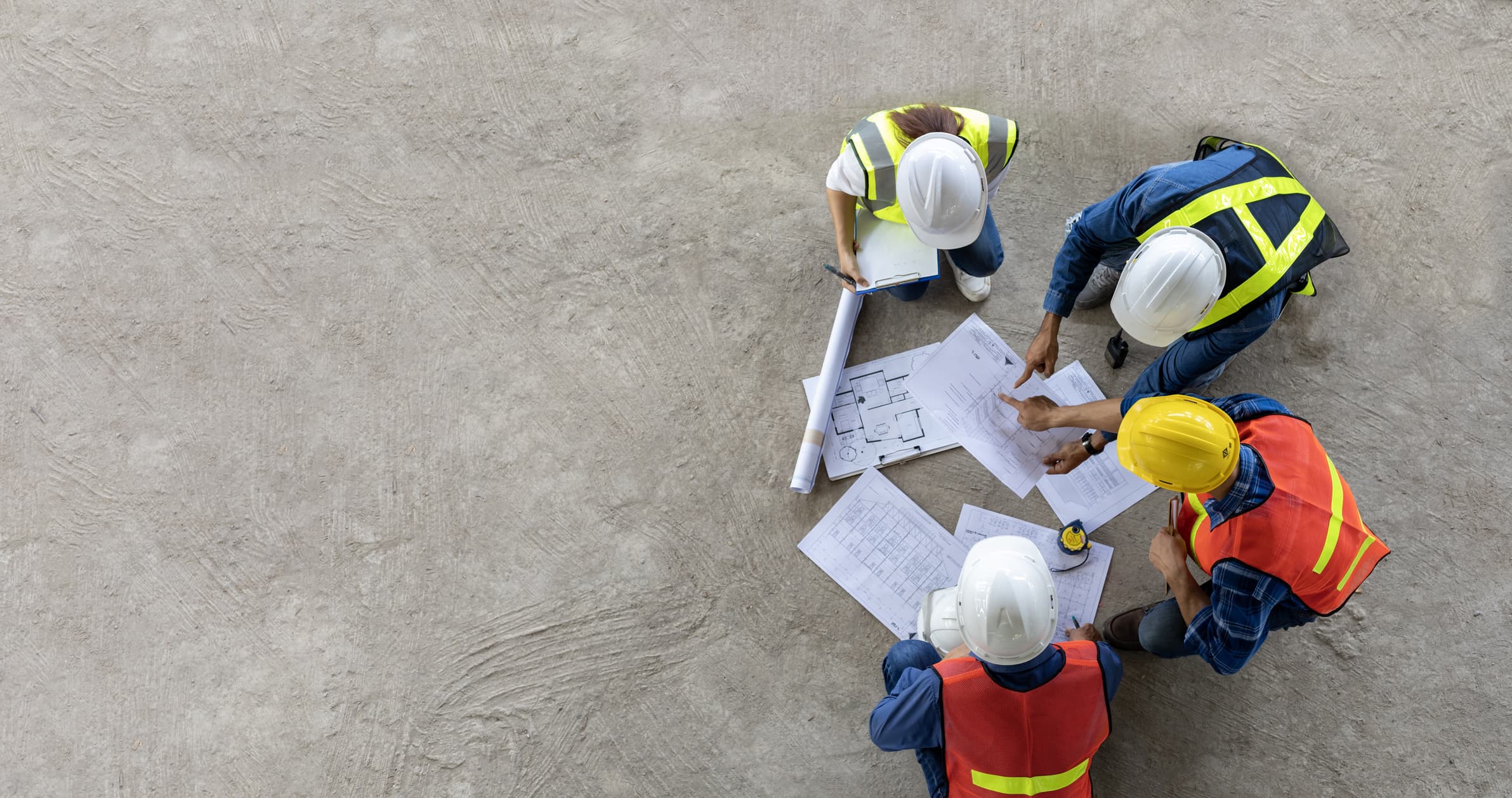Four people wearing hardhats and safety vests reviewing blueprints on a sandy surface