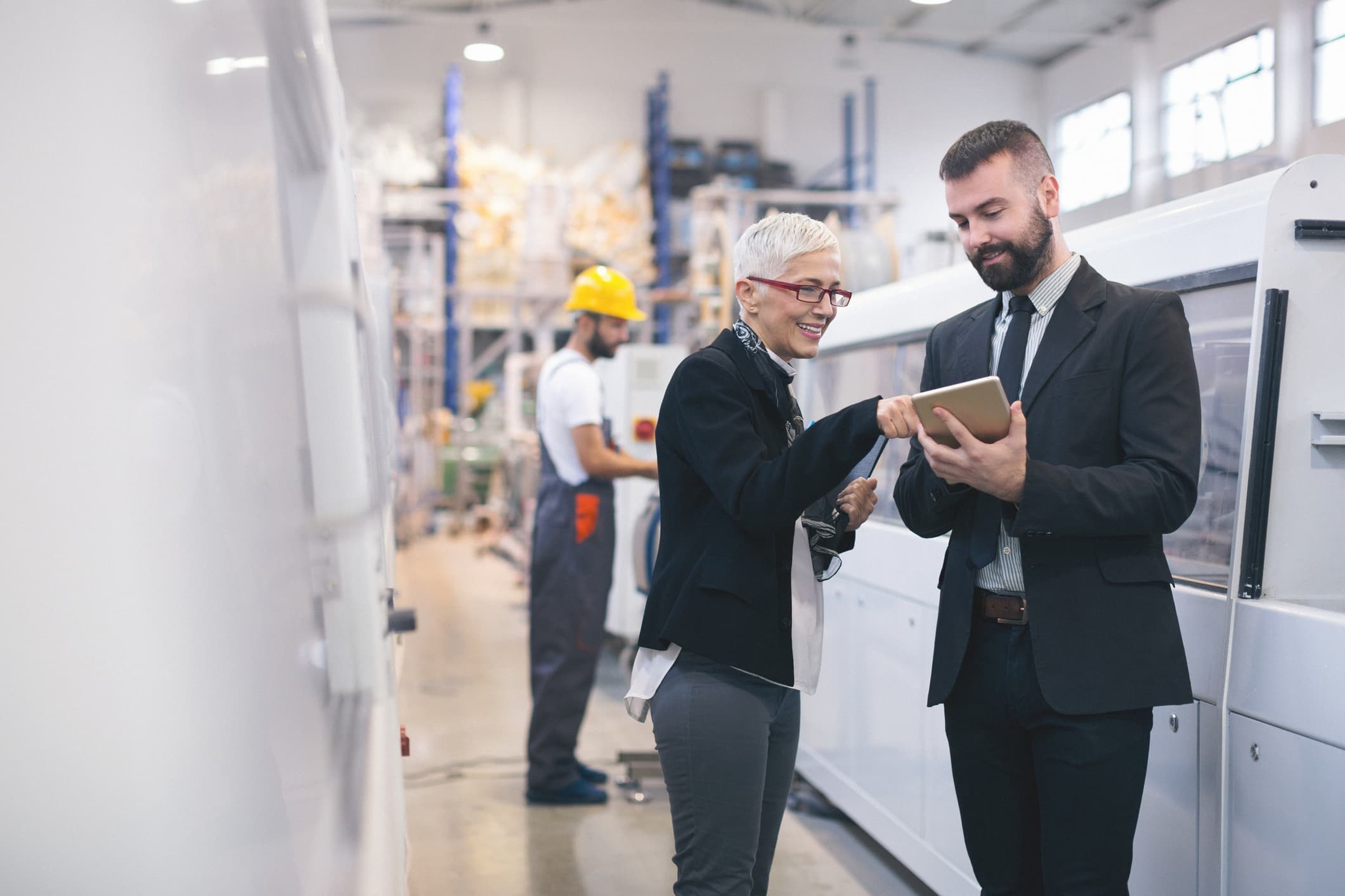 woman pointing at tablet in male colleagues hand in factory with man in hard hat in background